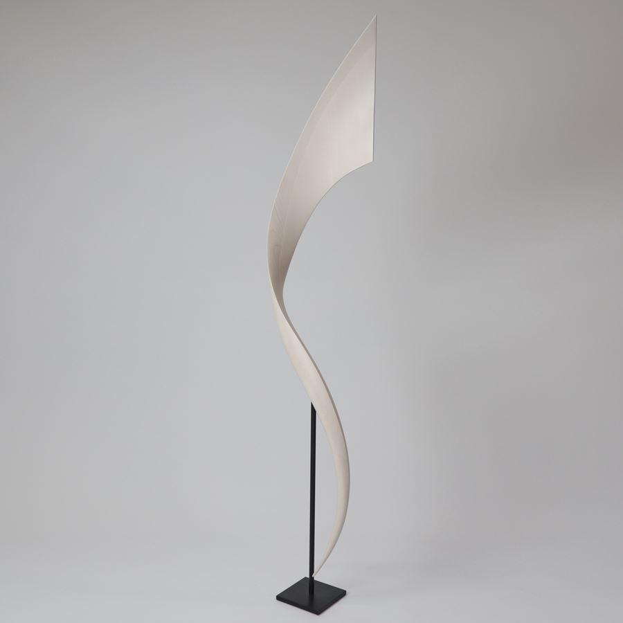 tall standing curved flaring white wooden sculpture with narrow base widening towards to the top and as it curves around presented on a black steel simple stand