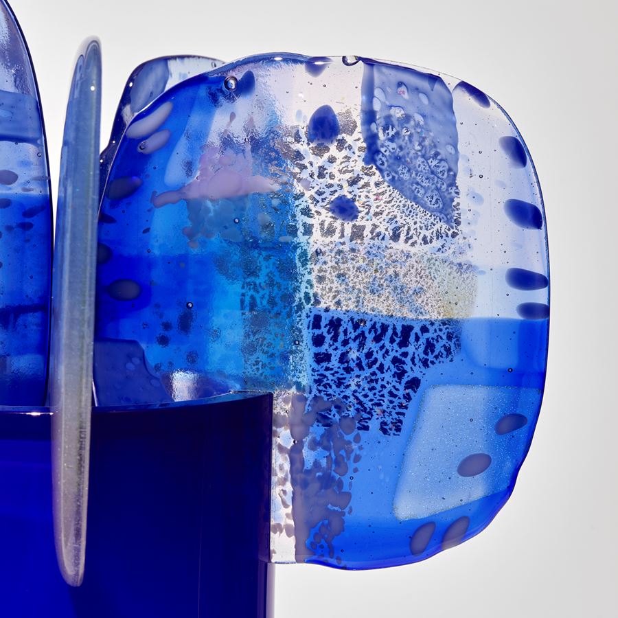 shiny blue cylinder fading from light to darker intensity of colour at the top with five petals perched on the top with abstract patterns and dots in blue and lilac hand made from blown and fused glass 