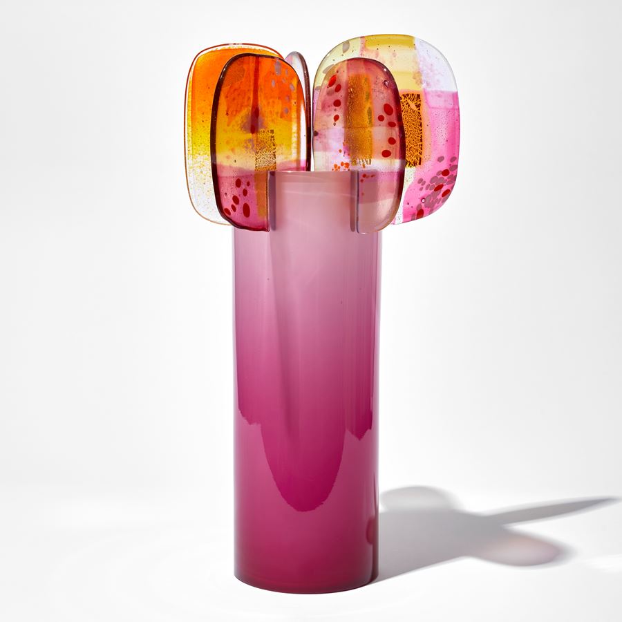 rich shiny opaque pink cylinder with colour fade towards the top with five finials overlapping the top edge each with abstract patterns in pink yellow and gold hand made from blown and fused glass