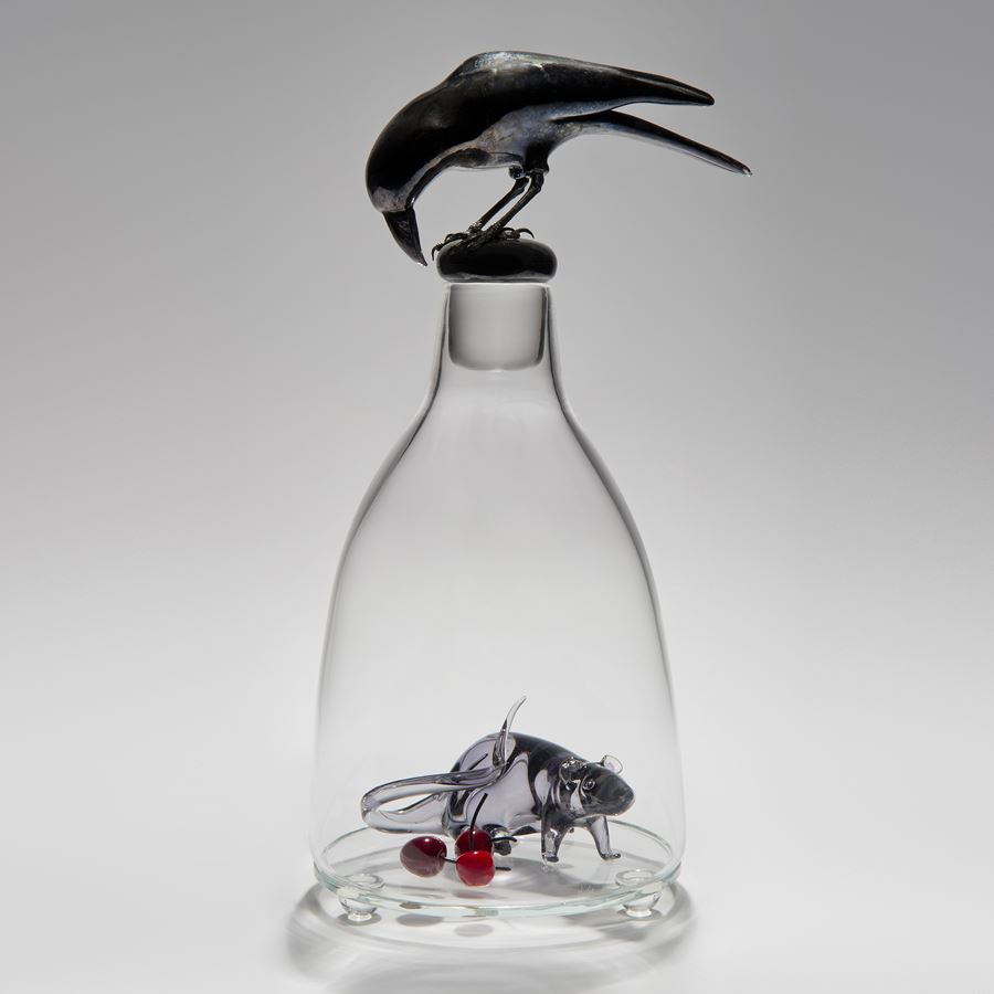 glass and steel sculpture of a mouse trapped in a clear glass flask with a crow peering in from the top
