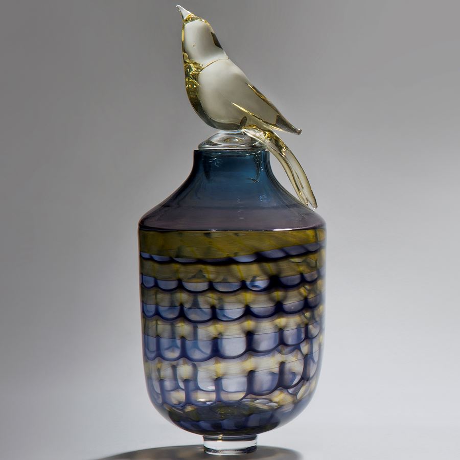 glass artwork of bird sat on top of funeral urn in blue purple yellow and gold