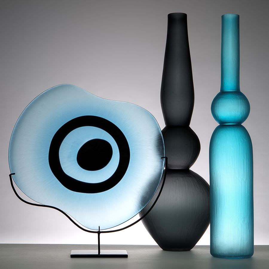 sculpted glass plate in light blue with black eye shape on metal stand