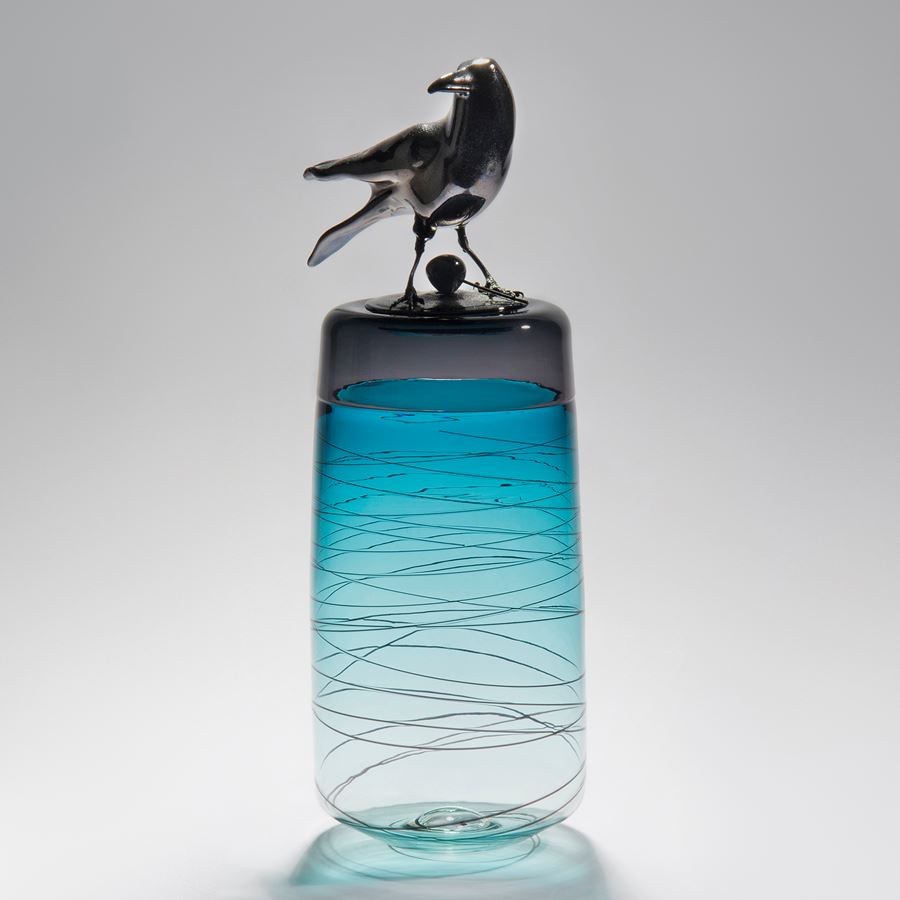 sculpted glass vase in clear and turquoise with faint lines encircling exterior with stainless steel crow on top
