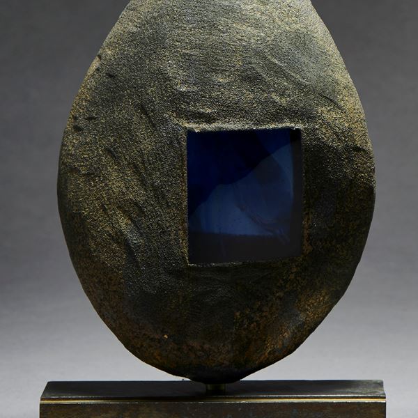 grey and brown hand made glass and metal ovoid shaped sculpture with square glass blue window and square geometric aged metal base