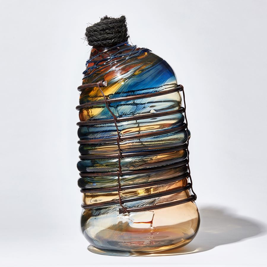 glass sculpture in the shape of an old bottle in blue clear and amber glass in a  copper cage with rope stopper