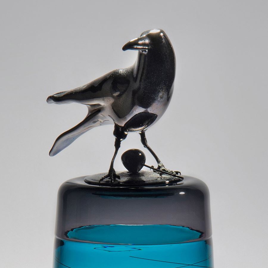 sculpted glass vase in clear and turquoise with faint lines encircling exterior with stainless steel crow on top