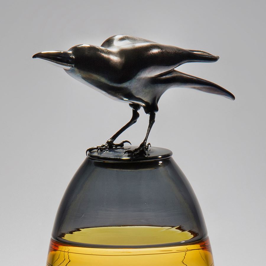 glass and steel sculpture of a crow atop a clear and yellow tinted vase with helter skelter pattern