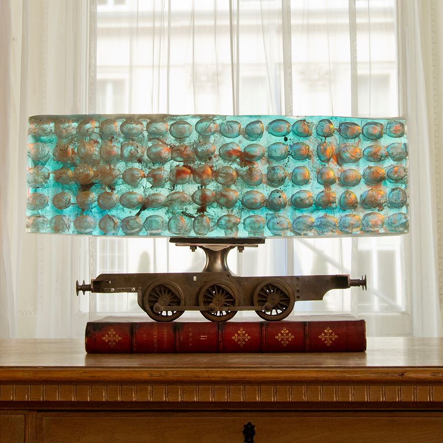 ethereal aqua and jade coloured large rectangular mass of glass with ovoid cutout sections perched on top of a metal scale model train chassis 