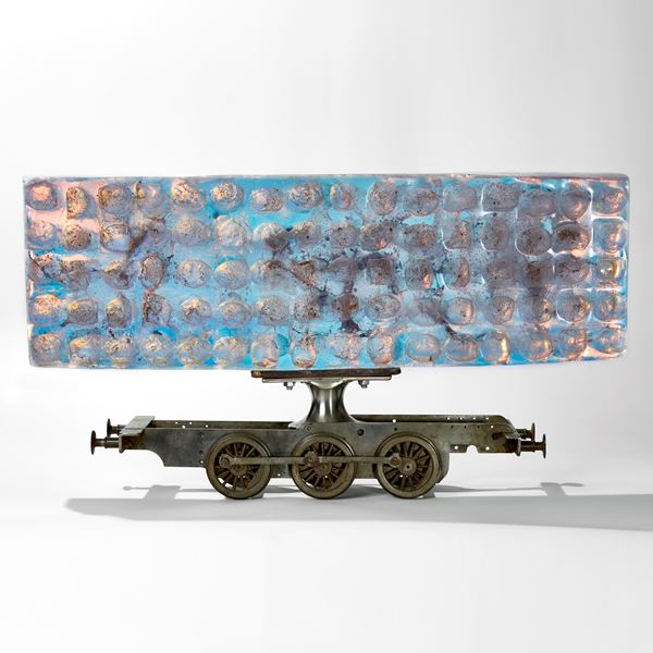 ethereal aqua and jade coloured large rectangular mass of glass with ovoid cutout sections perched on top of a metal scale model train chassis 