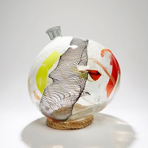 round bottle sculpture in clear with latticed black detail and patches of red yellow and white hand made from glass with ocean plastic fitting at the top