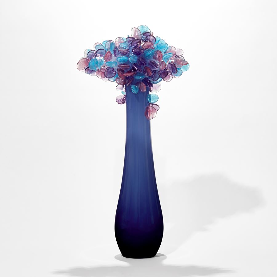simplified tree with elongated tear shaped smooth trunk in dark blue lightening to the top with a cluster of lollipop leaves on top in blue and lilac handmade from glass