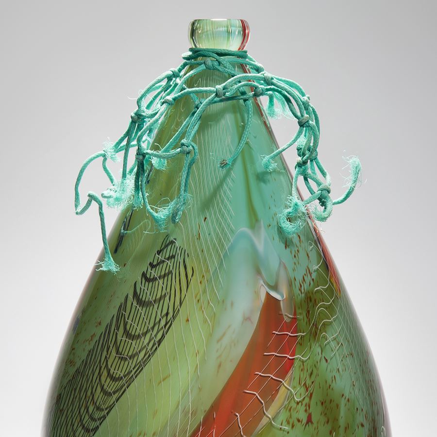 smooth droplet shaped green and reddish amber sculpture with lined areas of detail hand made from glass with ocean plastic cap and trim of found fishing net