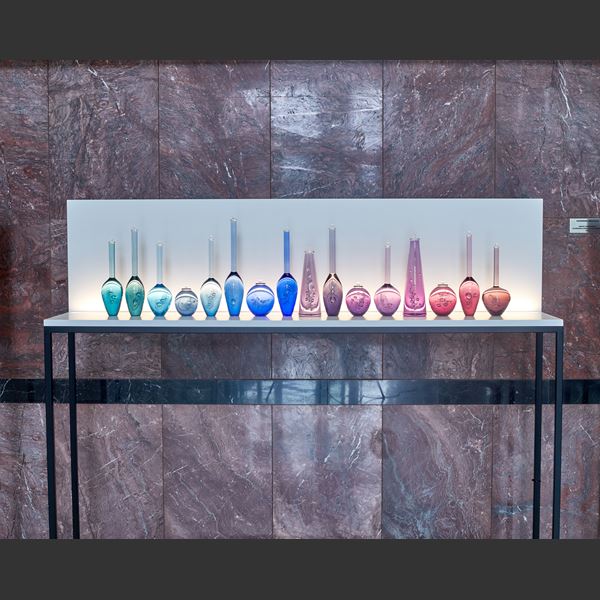 sixteen scientific looking solid bottles all different shapes each a different colour moving from aquas blues to pinks and rubies each hand made from glass