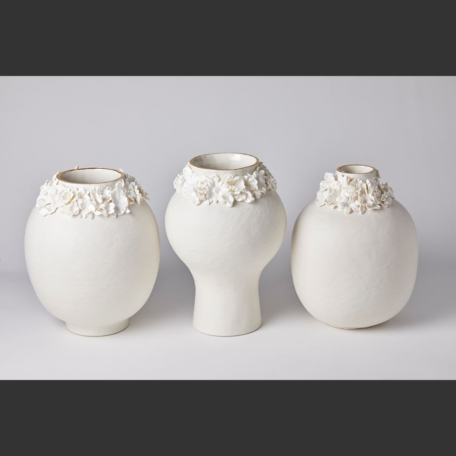 off white bulbous vase with narrow bottom half and slight rough texture with floral adornment around the neck which has a gold painted edge handmade from porcelain and parian