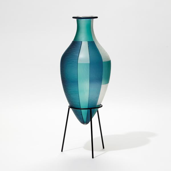blue jade and white elongated bottled with pointed base and wide rim and an abstract block cut pattern hand made from glass with a black steel stand