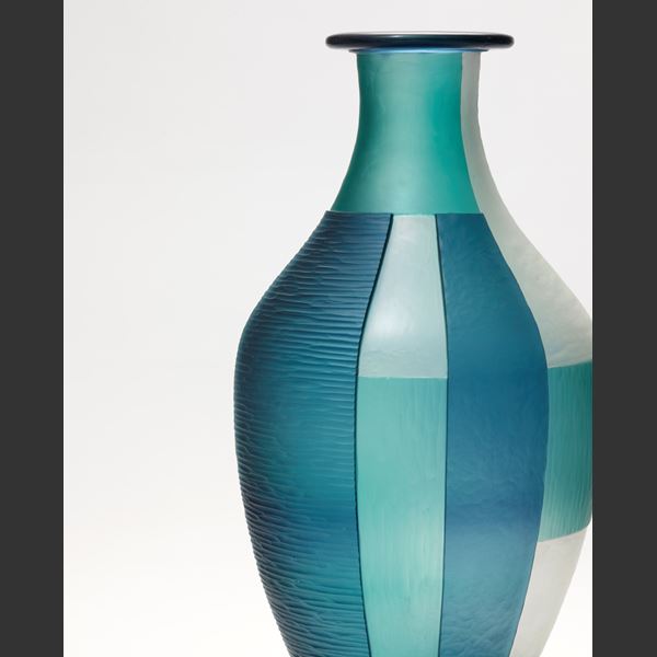 blue jade and white elongated bottled with pointed base and wide rim and an abstract block cut pattern hand made from glass with a black steel stand