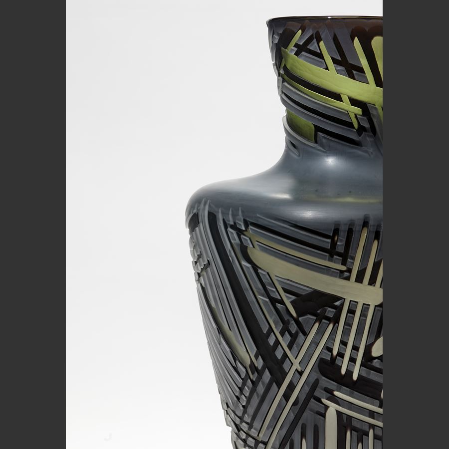 pointy bottomed bottle in dark grey and green which deep lines of differing widths cut in the surface hand made from glass with a steel stand 