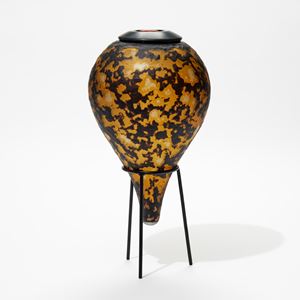 amber and black teardrop bottle with textured organic cut surface sat on a black tripod museum style stand