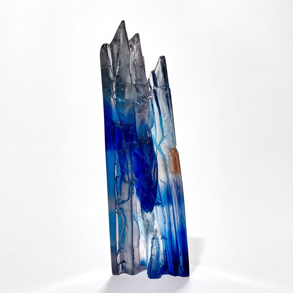 transparent angular sculpture with rugged cliff face detail with areas in light and dark blue hand made from cast glass