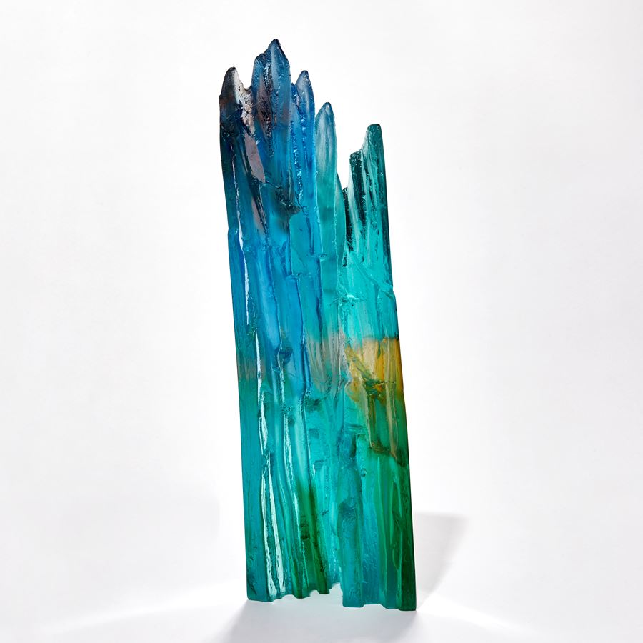 transparent abstract sculpture with two flat sheer sides and inner rugged cliff face textured edge in turquoise jade amber beige blue and red brown hand made from glass