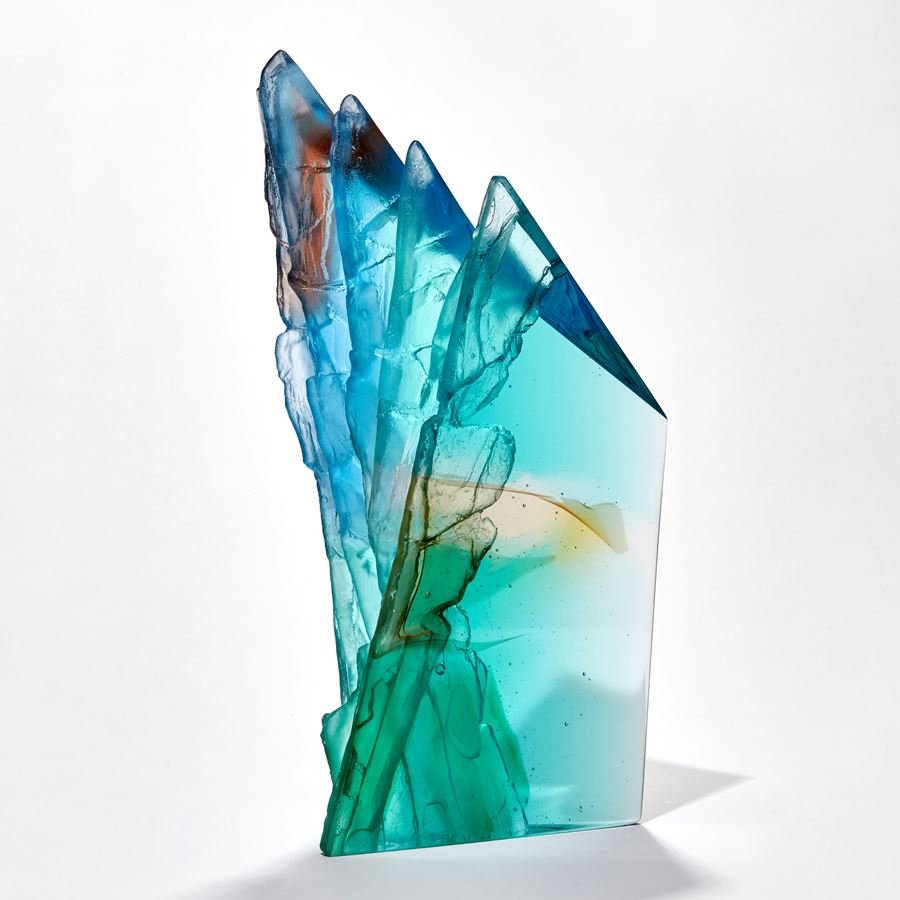 transparent abstract sculpture with two flat sheer sides and inner rugged cliff face textured edge in turquoise jade amber beige blue and red brown hand made from glass
