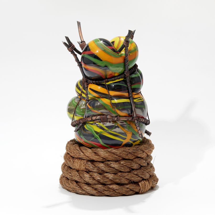 orange green yellow and black bulbous hand made glass sculpture held within a copper cage with a wounded rope at the base