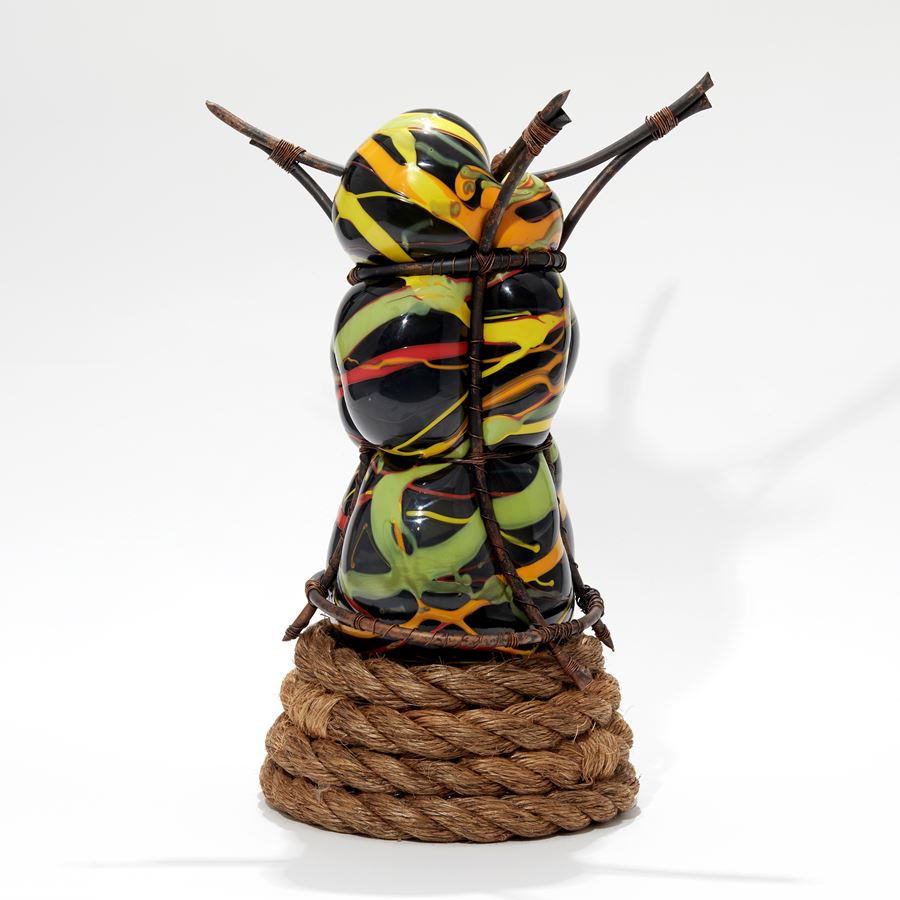 black yellow green red and orange undulating and bulbous handmade glass sculpture held within copper rods with wound rope base