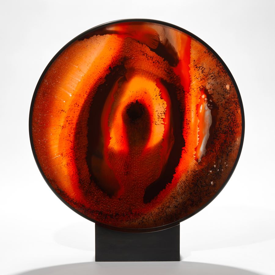 black round metal frame with solid rectangular base holding an abstract red orange and black glass artwork based on the evil amulet hand made from shattered and sheet glass