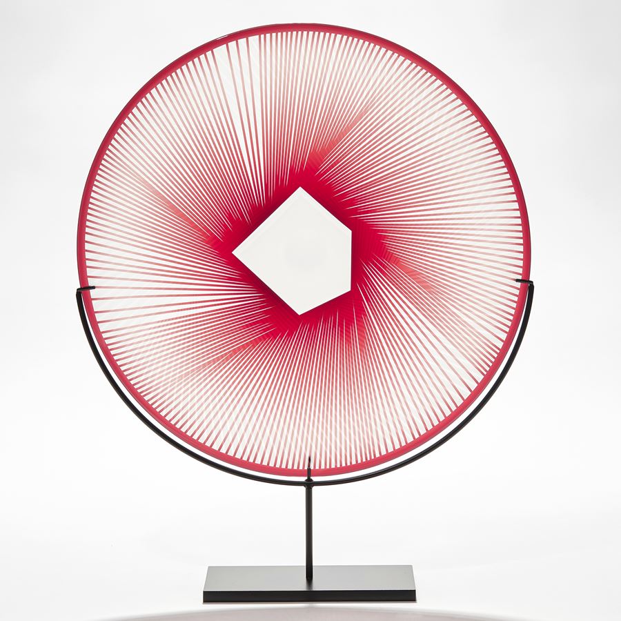 round glass plate on black metal stand with etched graphic pattern in clear and white with clear centre and red straight lines radiating from the middle