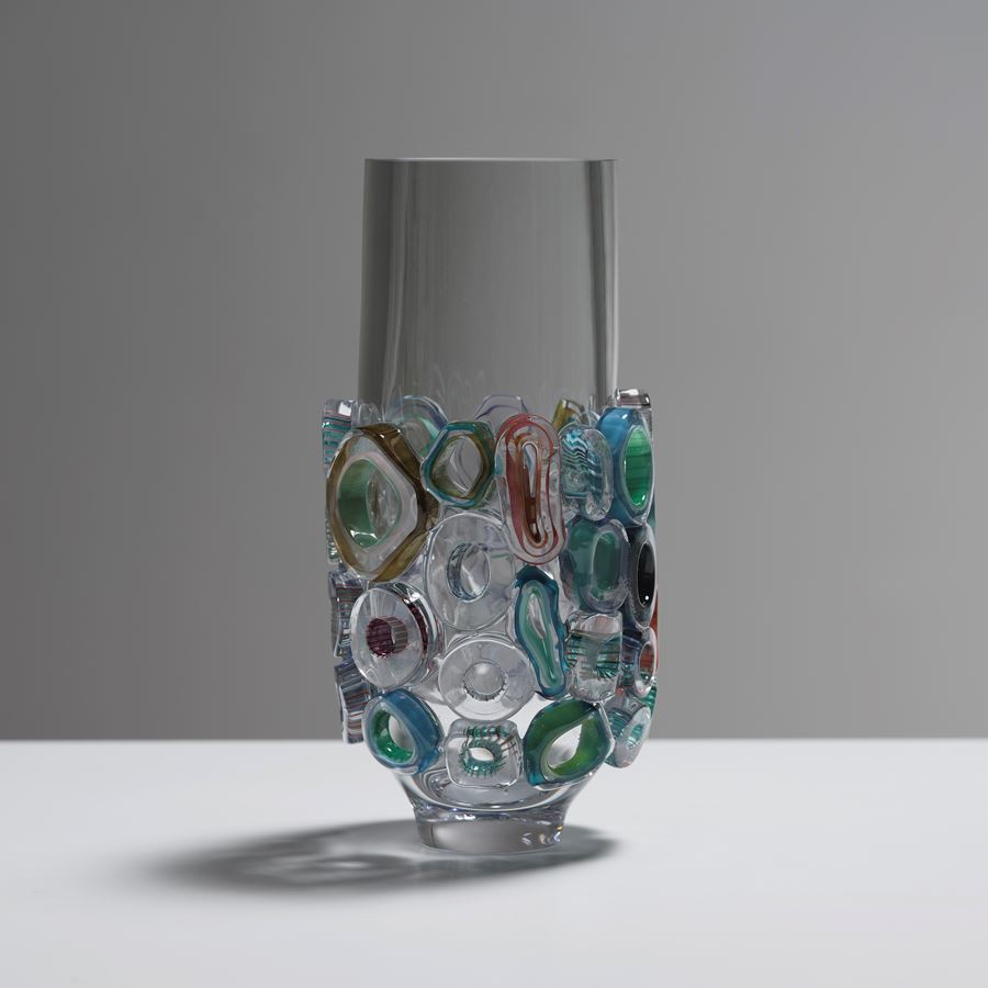 transparent tubular vase with lower half covered in raised sections in various shaped rings with different colours and patterns hand made from blown glass