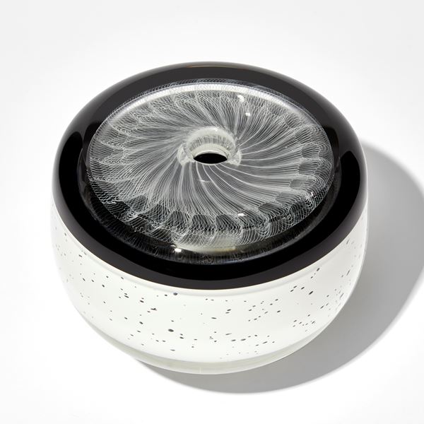 round vessel with small top opening in black and white with speckled base upper black band and white swirling patterning on the top flat surface hand made from glass