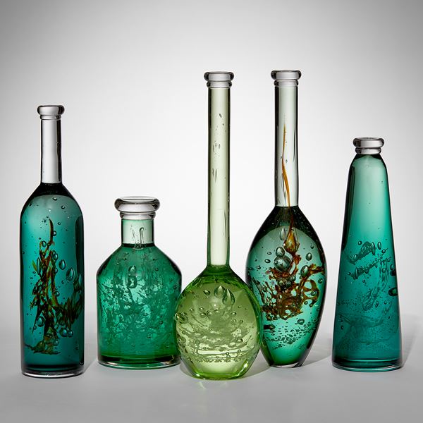 ten unique bottles of different shapes sizes and shades of green with mysterious organic trapped substances and details held within creating a unique installation hand made from glass