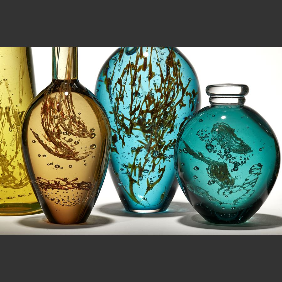 installation of seven solid transparent glass bottles in jade green ochre gold and khaki with organic plant life details trapped inside each with a rounded base and long thin neck and stopper hand made from glass