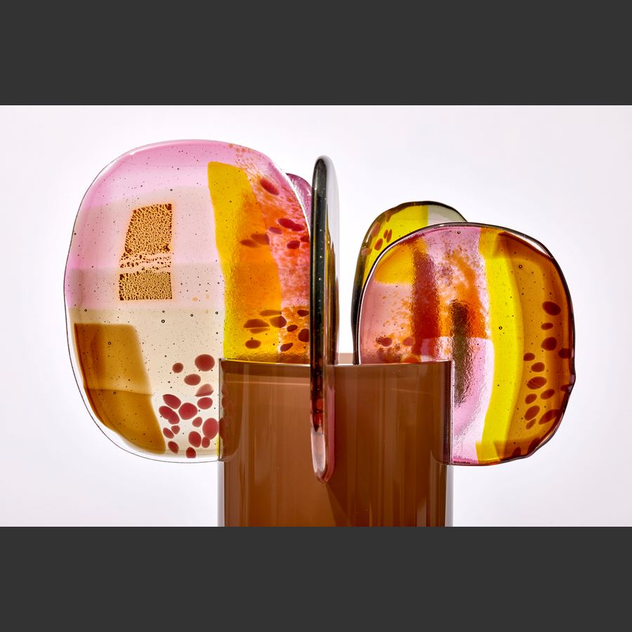 aubergine brown fading to the top opaque cylinder with five rounded panels of abstractly pattern glass in pink yellow gold and brown perched on the rim hand made from glass