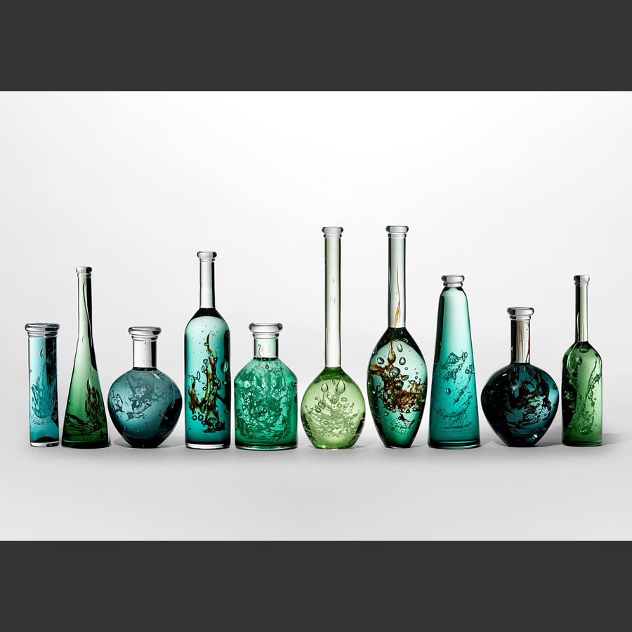 ten unique bottles of different shapes sizes and shades of green with mysterious organic trapped substances and details held within creating a unique installation hand made from glass