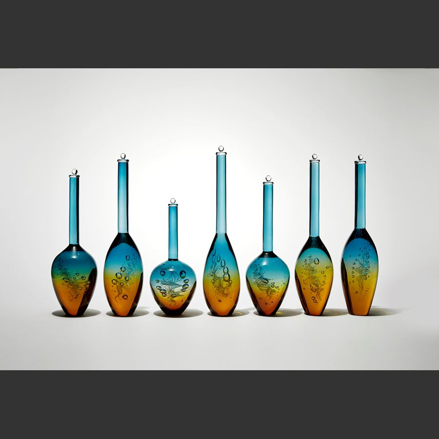 row of seven transparent solid bottles in different bulbous shapes with long thin necks with caps on top with amber bases merging to turquoise tops hand made from glass