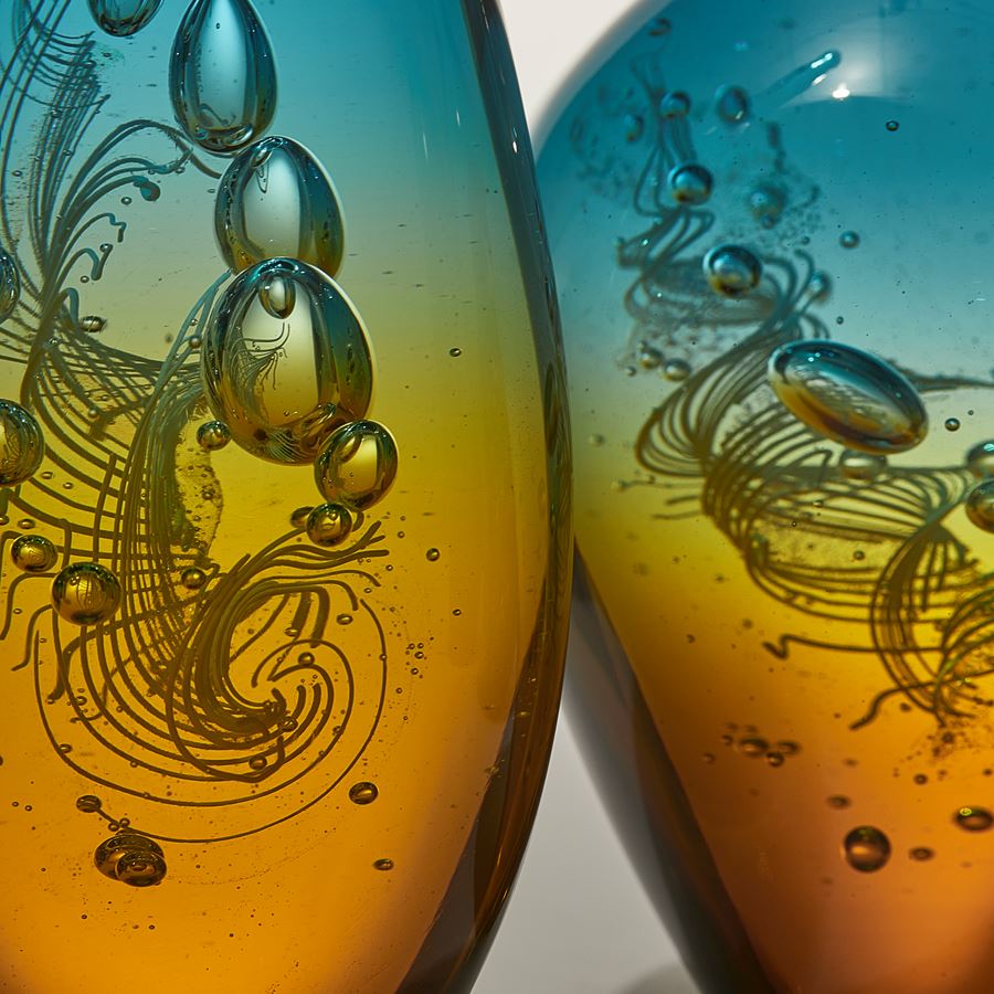 row of seven transparent solid bottles in different bulbous shapes with long thin necks with caps on top with amber bases merging to turquoise tops hand made from glass