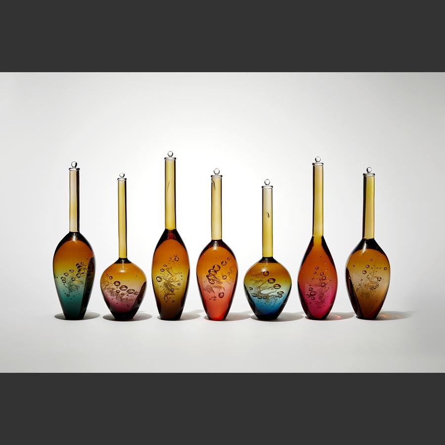 installation of seven transparent solid bottles in a row with spiral helix detail trapped within each with a long thin neck and bulbous base fading from amber at the top to various colours at the base hand made from glass