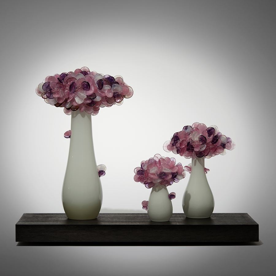 a trio of abstract trees each with an sleek alabaster coloured trunk topped with a multitude of sugary candy cane pink coloured lolliop leaves hand made from glass and presented on a rectangular wooden base