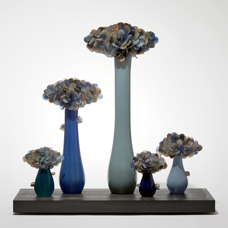 five simplified abstract trees with grey and dark blue trunks each densely covered at the top with lollipop shaped rounded leaves in bronze blue and grey hand made from glass with all sat on a wooden rectangular base 