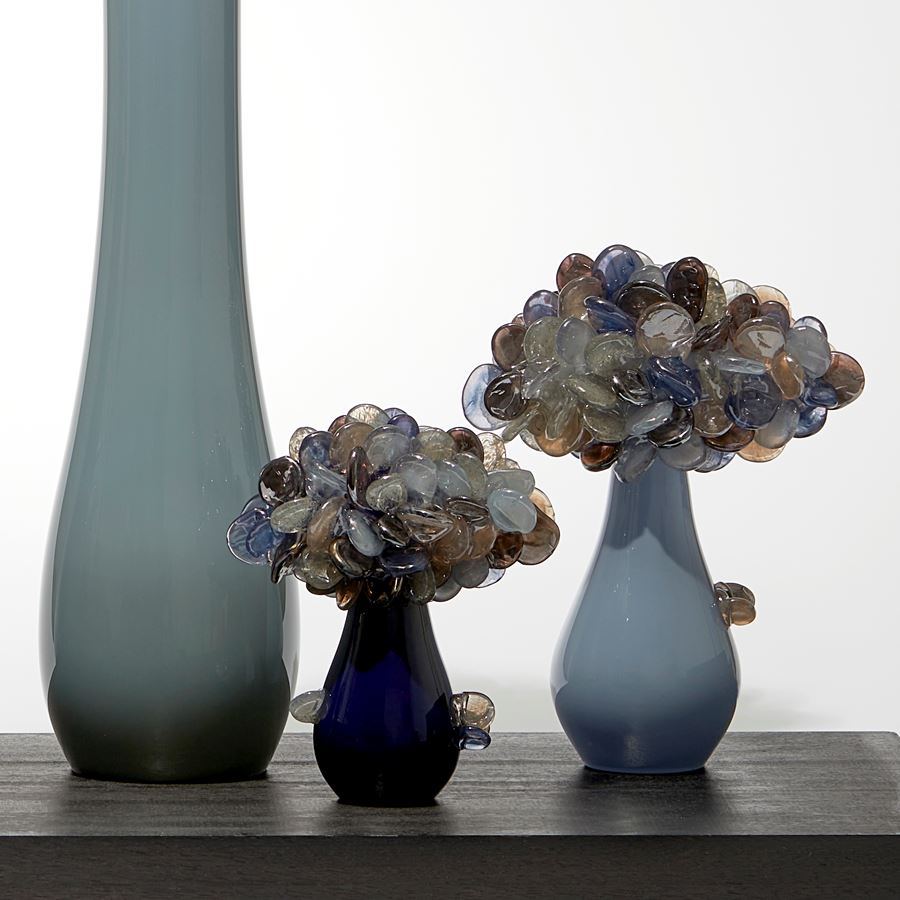 five simplified abstract trees with grey and dark blue trunks each densely covered at the top with lollipop shaped rounded leaves in bronze blue and grey hand made from glass with all sat on a wooden rectangular base 
