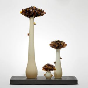 three trees with long sleek off white trunks topped with densely clustered rounded leaves in dark amber brown and grey hand made from glass and presented on a wooden base emulating bonsai