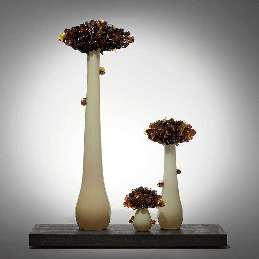 three trees with long sleek off white trunks topped with densely clustered rounded leaves in dark amber brown and grey hand made from glass and presented on a wooden base emulating bonsai