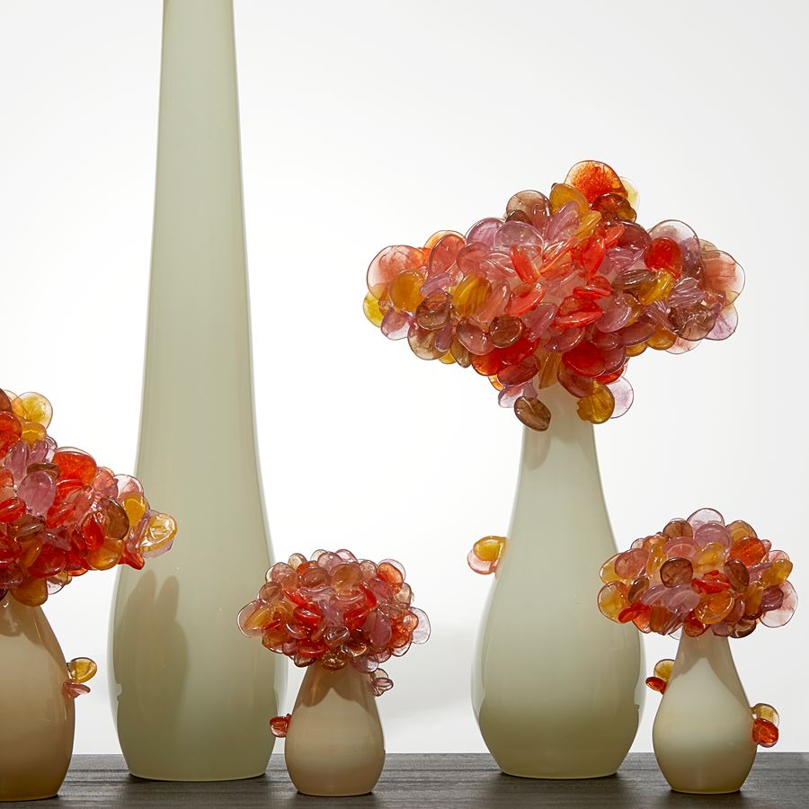 five abstract trees of differing heights with cream sleek trunks topped with clustered orange and amber lollipop shaped leaves hand made from glass and presented like bonsai on a rectangular wood base