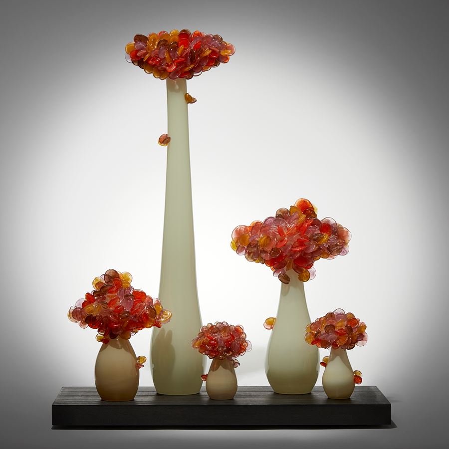 five abstract trees of differing heights with cream sleek trunks topped with clustered orange and amber lollipop shaped leaves hand made from glass and presented like bonsai on a rectangular wood base