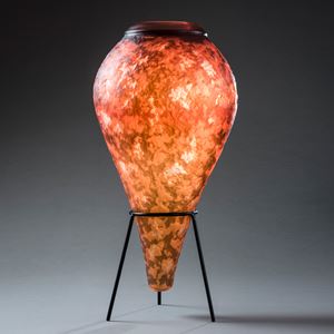 inverted teardrop shaped vessel with lava like textured mottled surface in fiery orange brown and yellow with top opening with a thick black ring hand made from glass sat upon a black steel tripod stand 
