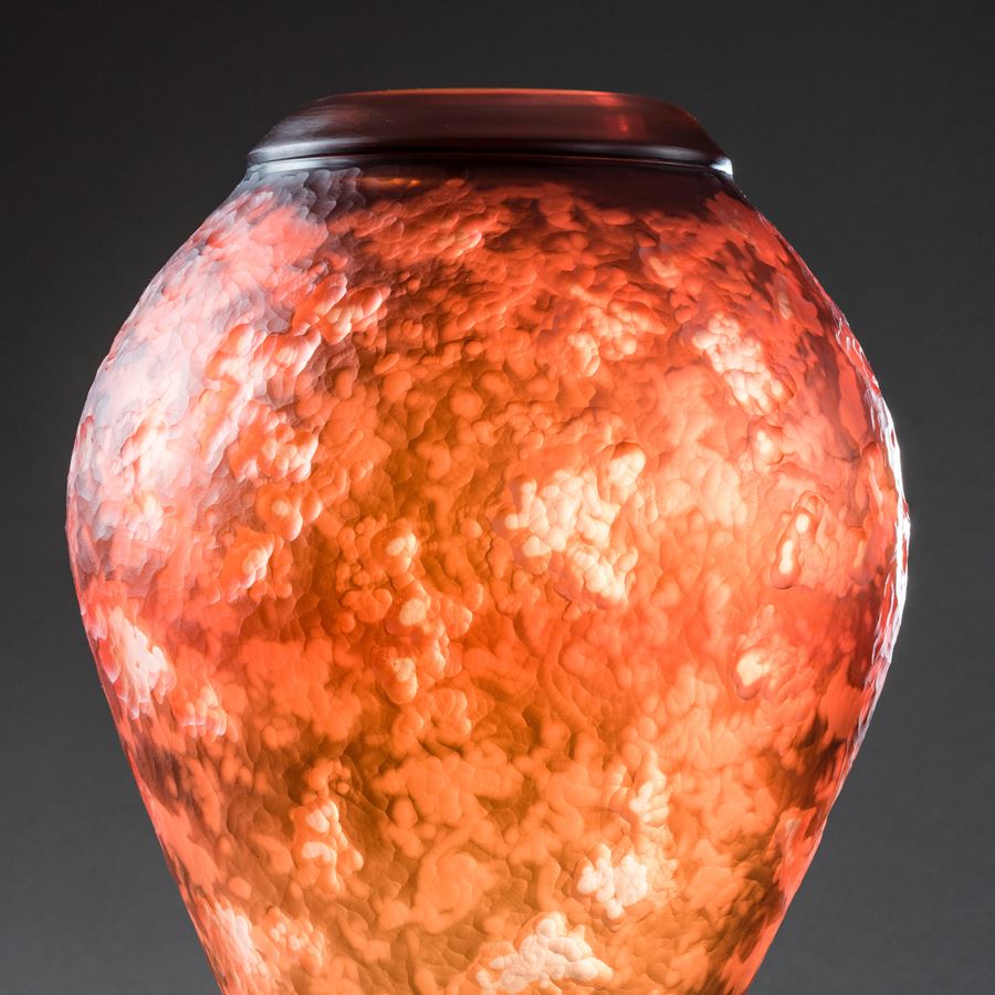 inverted teardrop shaped vessel with lava like textured mottled surface in fiery orange brown and yellow with top opening with a thick black ring hand made from glass sat upon a black steel tripod stand 