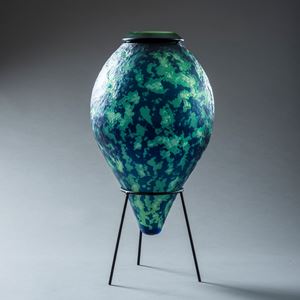 mottled organic textured rich dark blue and jade green inverted teardrop shaped vessel with top opening and think black rim hand made from glass and sat upon a black steel tripod stand