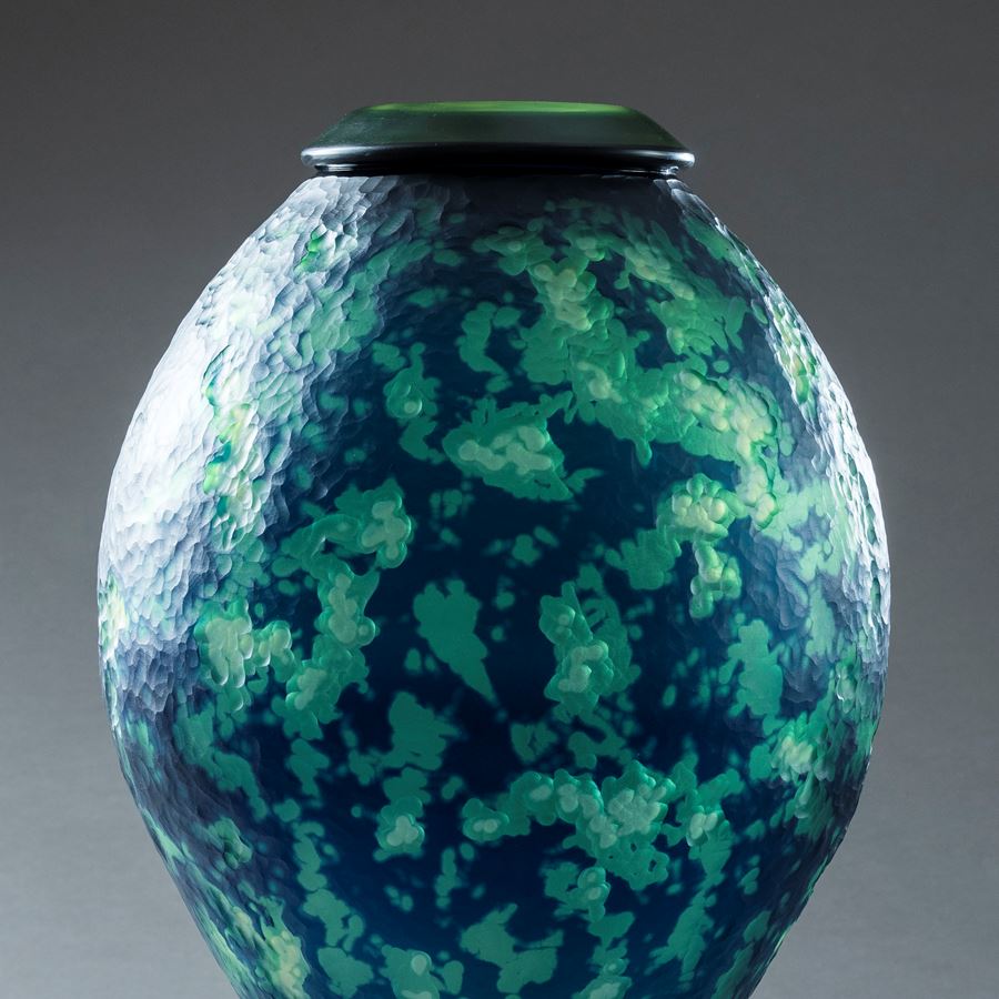 mottled organic textured rich dark blue and jade green inverted teardrop shaped vessel with top opening and think black rim hand made from glass and sat upon a black steel tripod stand