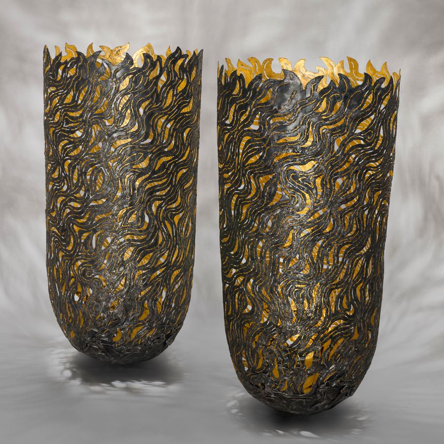tall darkened steel vessels with rounded bases and jagged flamed shaped top edges with arced rounded curling patterns on the surface and cut out sections with a gold reflective interior surface
